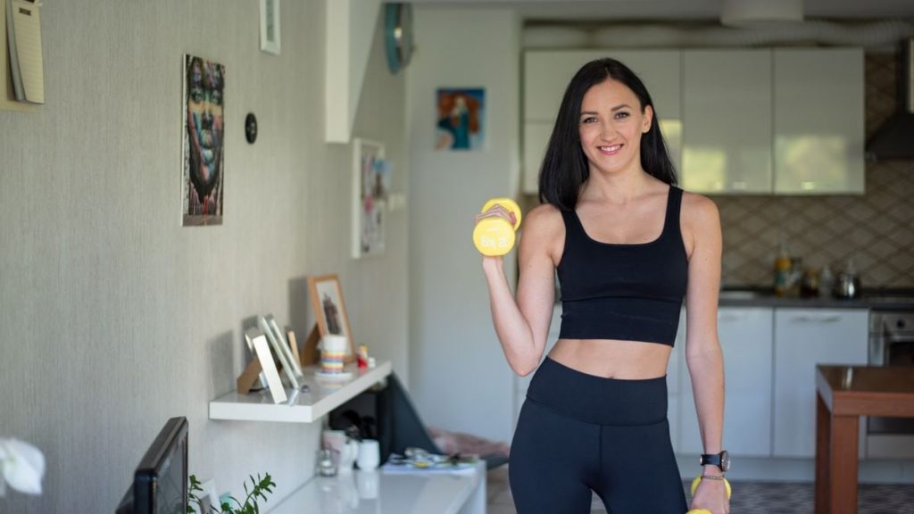 Smiling woman in activewear lifting dumbbells at home for a healthy lifestyle.